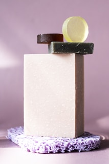 A set of soap bars arranged in a minimalistic style. A large rectangular off-white soap stands vertically, topped with smaller round soaps in green, brown, and yellow hues. The soaps rest on a textured lavender mat, against a softly lit lavender background.