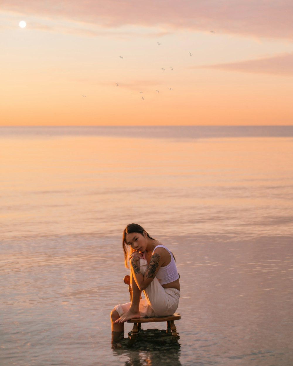 Femme en robe blanche assise sur une chaise en bois marron sur la plage au coucher du soleil