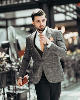 A well-dressed man in a grey plaid suit and orange tie is walking outdoors, holding a small brown leather accessory. He is adjusting his tie and looking ahead, exuding confidence. The background is blurred, suggesting an urban setting with plants on the side.