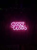Bright pink neon sign displaying the words 'Good Vibes' against a dark background with a subtle bamboo texture.