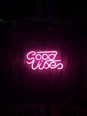 Bright pink neon sign displaying the words 'Good Vibes' against a dark background with a subtle bamboo texture.