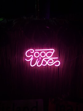 Bright pink neon sign displaying the words 'Good Vibes' against a dark background with a subtle bamboo texture.