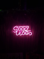 Bright pink neon sign displaying the words 'Good Vibes' against a dark background with a subtle bamboo texture.