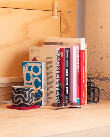 A wooden desk holds a collection of vertically stacked books between two black metal bookends. In front of the books is a ceramic mug with a wavy black and white pattern, placed on a red coaster. A pair of sunglasses rests nearby on the desk. The background features a light wooden wall and part of a concrete wall.