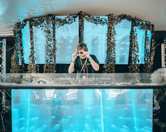 A DJ is performing on stage, adjusting their sunglasses. The setup includes a large mixing console and a backdrop of vibrant blue panels decorated with greenery. The scene has a modern and energetic atmosphere.