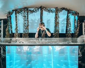 A DJ is performing on stage, adjusting their sunglasses. The setup includes a large mixing console and a backdrop of vibrant blue panels decorated with greenery. The scene has a modern and energetic atmosphere.