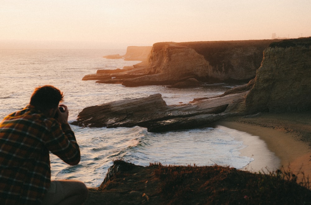 man in black and gray jacket sitting on rock formation near body of water during daytime