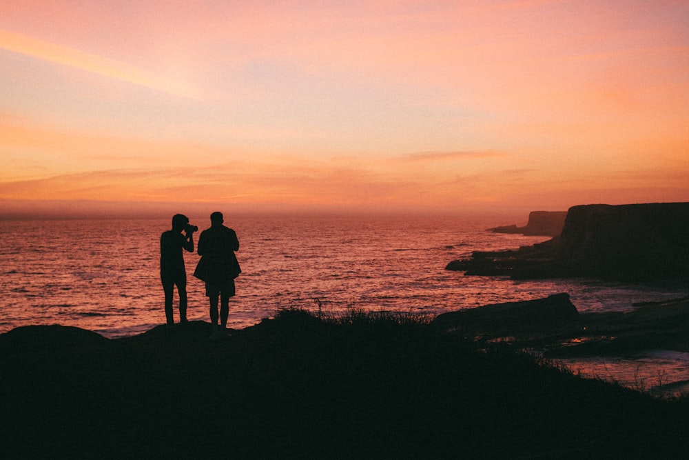 silhouette of 2 person standing on rock formation near body of water during sunset