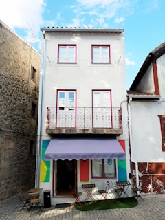 A three-story narrow building with a white facade and red-trimmed windows. The ground floor has a small entrance with a colorful awning and a few outdoor tables and chairs set on a cobblestone street. The building is flanked by older stone structures, and there is a clear blue sky above.