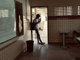 A person wearing a backpack stands in the doorway of a classroom, looking at a phone. The room has tiled walls, a whiteboard, and desks. Outside, there is a courtyard area with trees visible in the background.