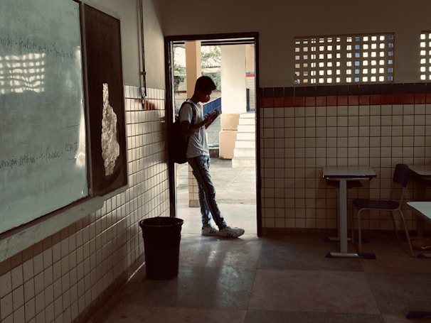 A person wearing a backpack stands in the doorway of a classroom, looking at a phone. The room has tiled walls, a whiteboard, and desks. Outside, there is a courtyard area with trees visible in the background.