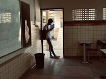 A person wearing a backpack stands in the doorway of a classroom, looking at a phone. The room has tiled walls, a whiteboard, and desks. Outside, there is a courtyard area with trees visible in the background.