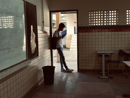 A person wearing a backpack stands in the doorway of a classroom, looking at a phone. The room has tiled walls, a whiteboard, and desks. Outside, there is a courtyard area with trees visible in the background.