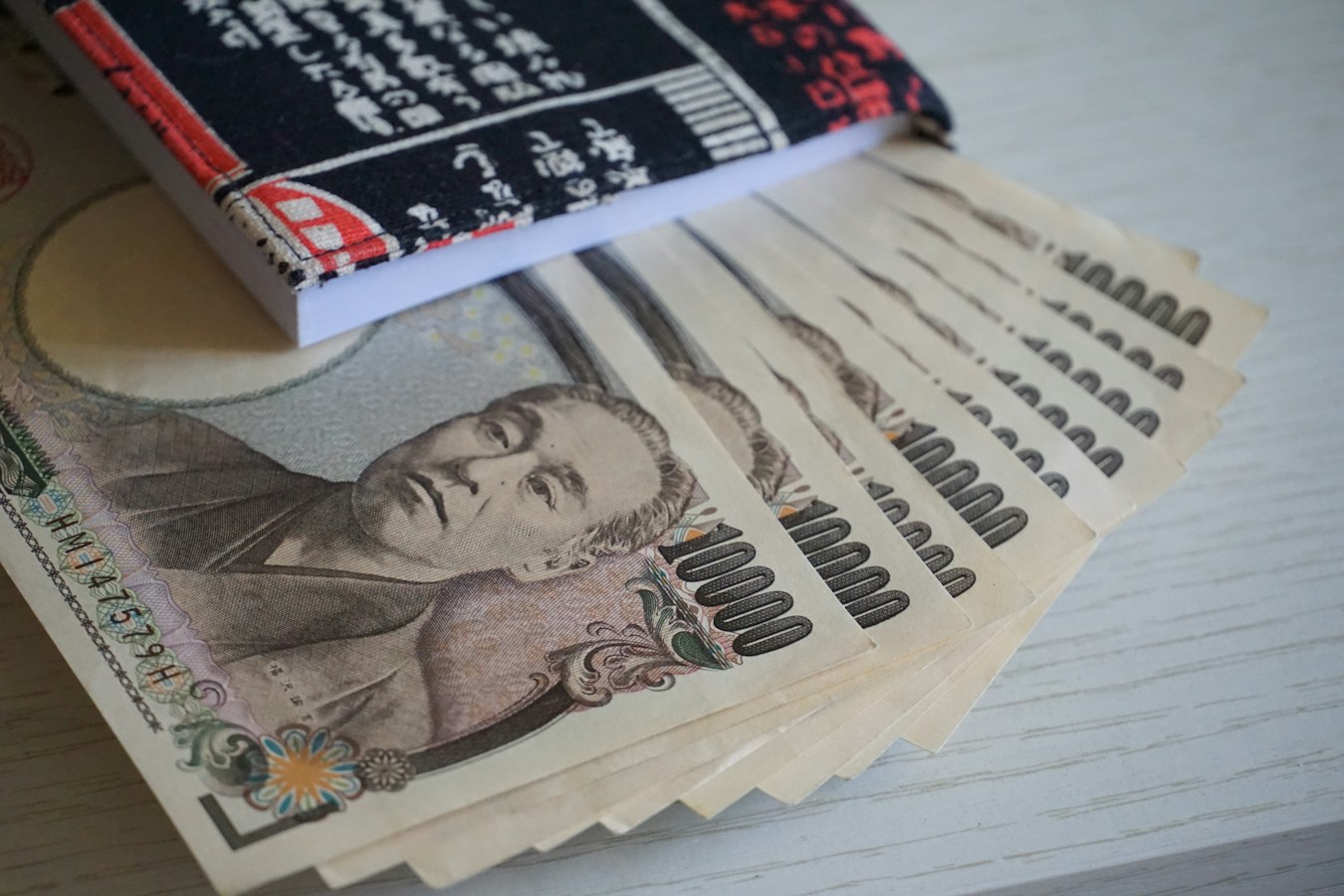 A fanned-out stack of Japanese 10,000-yen banknotes bearing the Yukichi Fukuzawa portrait, laid on a pale wooden table beside a black-and-red