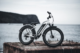A sleek, white electric bicycle with black accents is stationed on a weathered concrete slab overlooking a calm body of water. The scene includes a distant view of a rocky, tree-covered hill shrouded in mist.