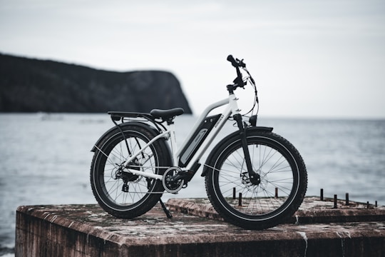 A sleek, white electric bicycle with black accents is stationed on a weathered concrete slab overlooking a calm body of water. The scene includes a distant view of a rocky, tree-covered hill shrouded in mist.