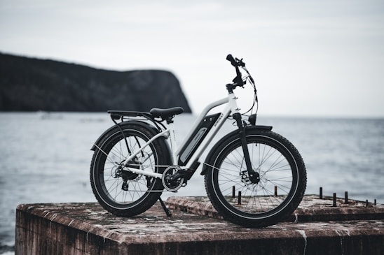 A sleek, white electric bicycle with black accents is stationed on a weathered concrete slab overlooking a calm body of water. The scene includes a distant view of a rocky, tree-covered hill shrouded in mist.
