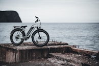 A white electric bicycle is parked on a weathered concrete pier facing the open sea. The background features an overcast sky and the distant silhouette of a rocky cliff. The setting gives a sense of solitude and adventure.