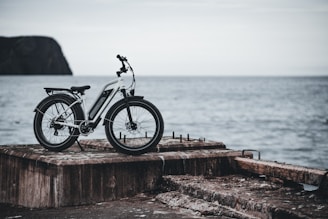 A white electric bicycle is parked on a weathered concrete pier facing the open sea. The background features an overcast sky and the distant silhouette of a rocky cliff. The setting gives a sense of solitude and adventure.