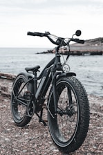 A black electric bike with thick, rugged tires is parked near a body of water, likely a sea or ocean. The bike is positioned on a rocky or gravelly surface, with a distant shoreline and cloudy sky in the background.