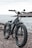 A black electric bike with thick, rugged tires is parked near a body of water, likely a sea or ocean. The bike is positioned on a rocky or gravelly surface, with a distant shoreline and cloudy sky in the background.