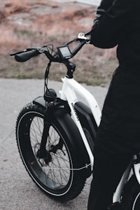 A person stands next to an electric bicycle with thick tires and a white frame. The handlebars feature a digital display, and the setting appears to be an outdoor area with a path and grass in the background.