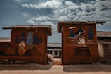 Two walls with rustic paintings depict scenes with human figures and abstract shapes against a backdrop of a rural setting. The sky is partially cloudy, adding depth and contrast to the earthy tones of the walls and the surrounding structures.