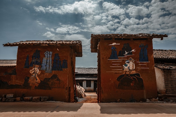 Two walls with rustic paintings depict scenes with human figures and abstract shapes against a backdrop of a rural setting. The sky is partially cloudy, adding depth and contrast to the earthy tones of the walls and the surrounding structures.