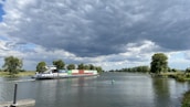 A large cargo ship travels along a river, carrying multiple colorful containers. There are green trees lining the riverbanks and a buoy floating in the water. The sky above is mostly covered with dark, thick clouds, suggesting the possibility of an approaching storm.