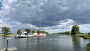 A large cargo ship travels along a river, carrying multiple colorful containers. There are green trees lining the riverbanks and a buoy floating in the water. The sky above is mostly covered with dark, thick clouds, suggesting the possibility of an approaching storm.