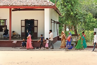 A group of people in traditional colorful attire, including women and children, walk past a single-story building with a tiled roof surrounded by green vegetation. Some individuals are seated inside the building with an open entrance. The scene appears to be a mix of activity and leisure.