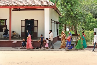 A group of people in traditional colorful attire, including women and children, walk past a single-story building with a tiled roof surrounded by green vegetation. Some individuals are seated inside the building with an open entrance. The scene appears to be a mix of activity and leisure.