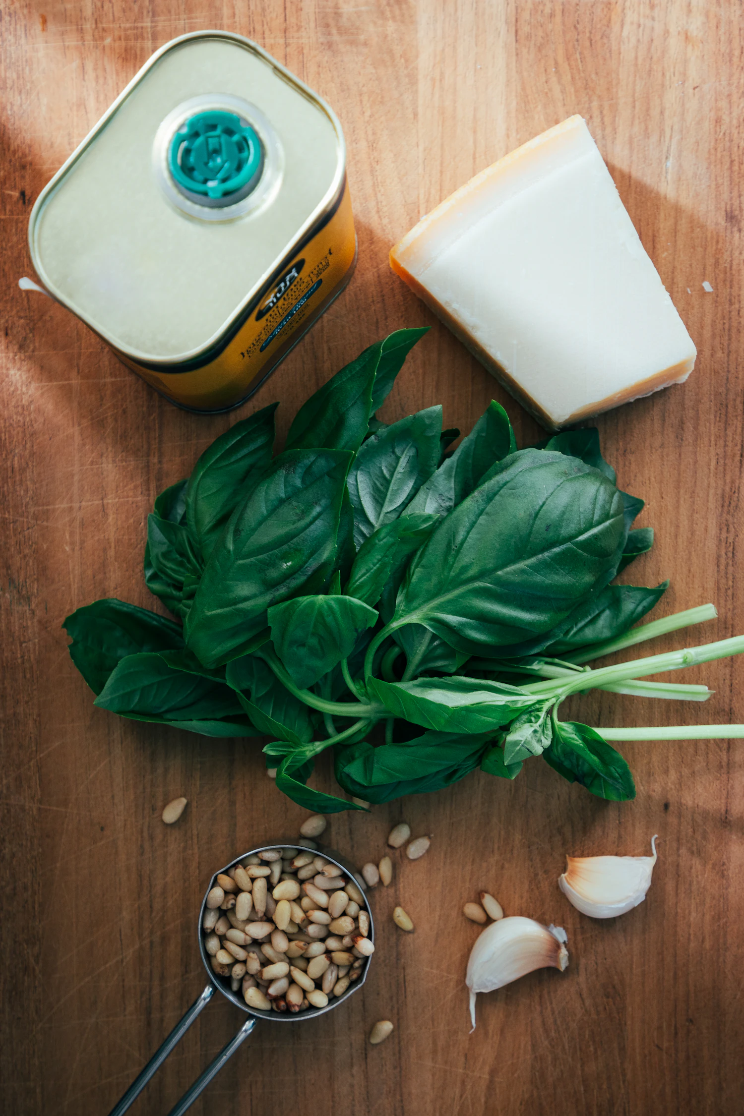 A bunch of fresh basil beside a tin of olive oil, a wedge of hard cheese and garlic on a wooden board