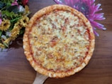 A freshly baked pizza with a golden-brown crust topped with melted cheese and bits of savory ingredients. It is placed on a wooden surface, accompanied by a colorful arrangement of yellow and pink flowers.