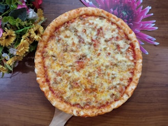 A freshly baked pizza with a golden-brown crust topped with melted cheese and bits of savory ingredients. It is placed on a wooden surface, accompanied by a colorful arrangement of yellow and pink flowers.