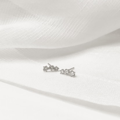 Two small, delicate stud earrings with multiple sparkling stones, placed on a textured white fabric background.
