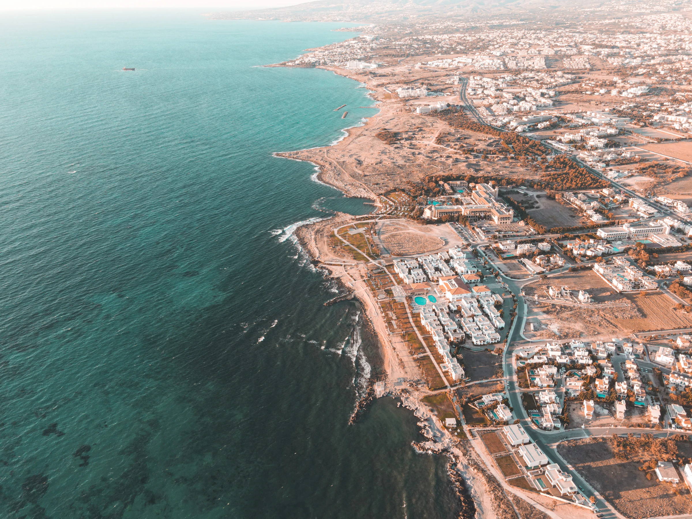 Aerial view of a Cypriot coastal resort, low white buildings running back from a rocky shoreline and turquoise sea
