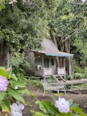 A rustic wooden cabin is nestled among lush greenery, surrounded by large trees and vibrant plants. The cabin features a small porch with a chair, and a canopy of hanging lights adds a charming touch. In the foreground, a garden bench and flowering plants provide a cozy and serene atmosphere.
