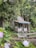 A rustic wooden cabin is nestled among lush greenery, surrounded by large trees and vibrant plants. The cabin features a small porch with a chair, and a canopy of hanging lights adds a charming touch. In the foreground, a garden bench and flowering plants provide a cozy and serene atmosphere.