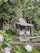 A rustic wooden cabin is nestled among lush greenery, surrounded by large trees and vibrant plants. The cabin features a small porch with a chair, and a canopy of hanging lights adds a charming touch. In the foreground, a garden bench and flowering plants provide a cozy and serene atmosphere.