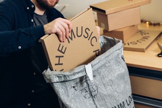 A person is placing a cardboard box labeled 'THE MUSIC P' into a large woven bag positioned on a wooden table. Several other similar boxes are stacked nearby. The individual is wearing a dark shirt, and the scene looks like a packing or organizing activity.