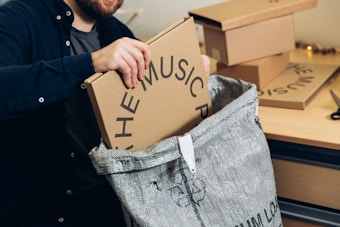 A person is placing a cardboard box labeled 'THE MUSIC P' into a large woven bag positioned on a wooden table. Several other similar boxes are stacked nearby. The individual is wearing a dark shirt, and the scene looks like a packing or organizing activity.