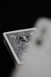 A deck of playing cards with an ornate, detailed, monochrome design is partially visible. The focus is on the intricate patterns and figures on the rear of the card. The background is dark, creating contrast against the light card.