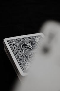 A deck of playing cards with an ornate, detailed, monochrome design is partially visible. The focus is on the intricate patterns and figures on the rear of the card. The background is dark, creating contrast against the light card.