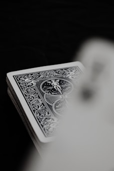 A deck of playing cards with an ornate, detailed, monochrome design is partially visible. The focus is on the intricate patterns and figures on the rear of the card. The background is dark, creating contrast against the light card.