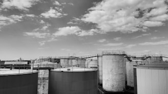 A series of large, cylindrical industrial tanks under a partly cloudy sky. The tanks are interconnected with metal walkways and ladders, suggesting an industrial facility setup.