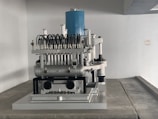 A complex mechanical apparatus with several metallic components, including pipes and cylinders, is prominently displayed. The machine features a blue cylindrical section at the top and is situated in a minimalist white room.