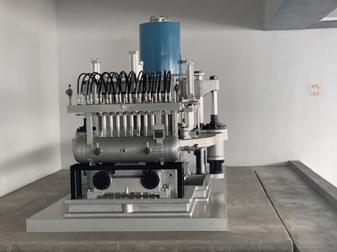 A complex mechanical apparatus with several metallic components, including pipes and cylinders, is prominently displayed. The machine features a blue cylindrical section at the top and is situated in a minimalist white room.