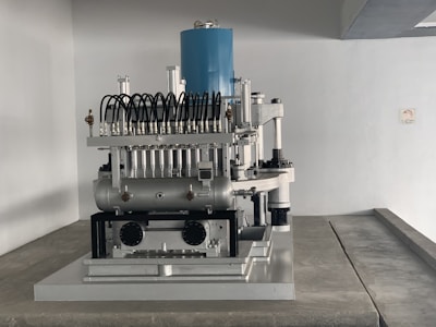 A complex mechanical apparatus with several metallic components, including pipes and cylinders, is prominently displayed. The machine features a blue cylindrical section at the top and is situated in a minimalist white room.