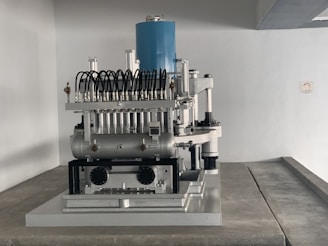 A complex mechanical apparatus with several metallic components, including pipes and cylinders, is prominently displayed. The machine features a blue cylindrical section at the top and is situated in a minimalist white room.